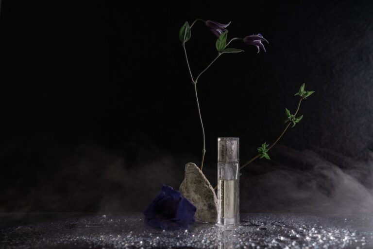 Artistic still life featuring purple flowers, rock, and glass perfume bottle against black background.