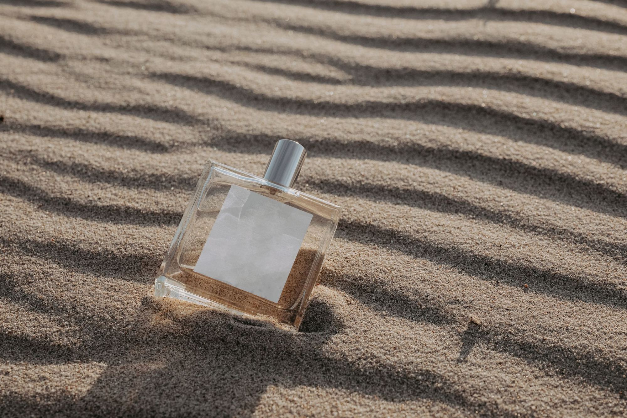 Stylish perfume bottle resting on rippled sand under sunlight, capturing a luxurious and serene atmosphere.